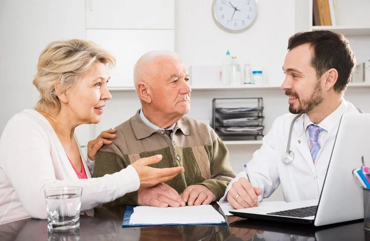 Doctor consulting with two older adults at desk in clinical setting.