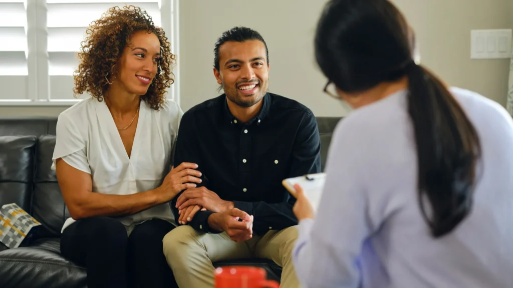 Two people consulting with advisor holding clipboard in an office setting.